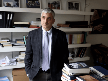 Man in suit stands in office with hands in his pockets.