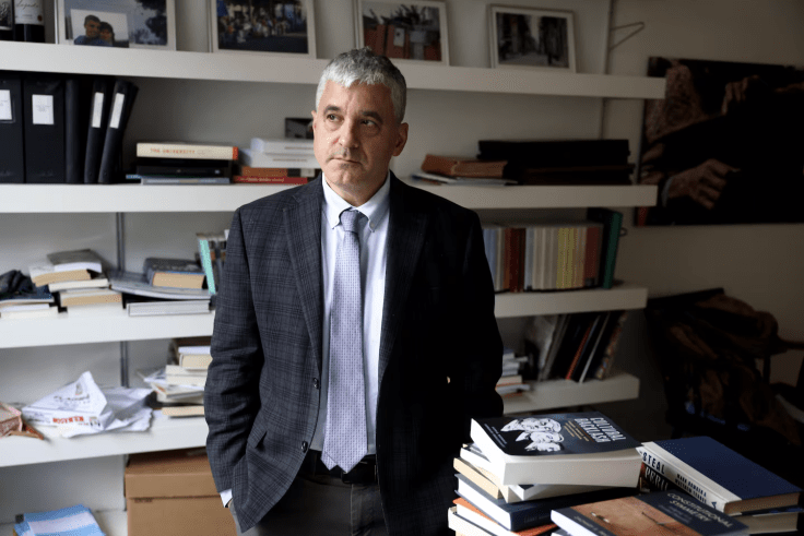 Man in suit stands in office with hands in his pockets.