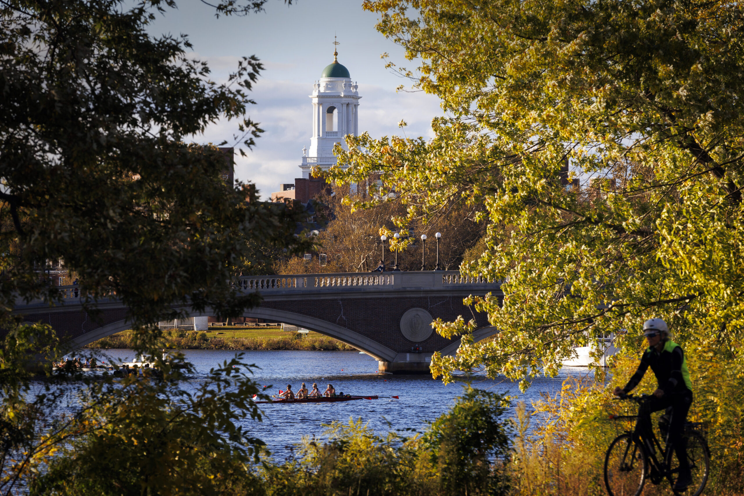 The peak of a Harvard building seen beyond the Charles river and behind trees.