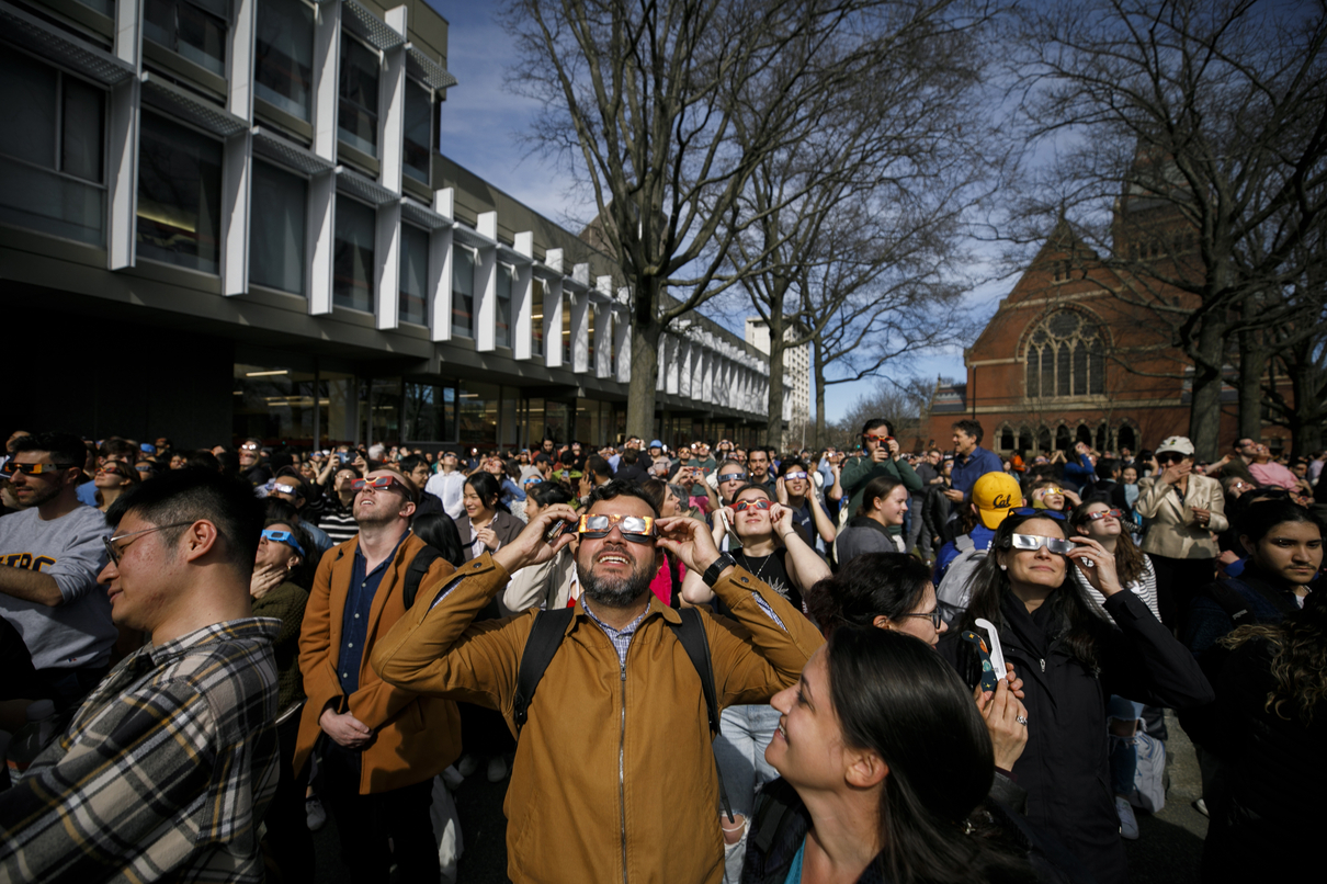 A man wearing glasses in a large crowd looks skyward at a solar eclipse.