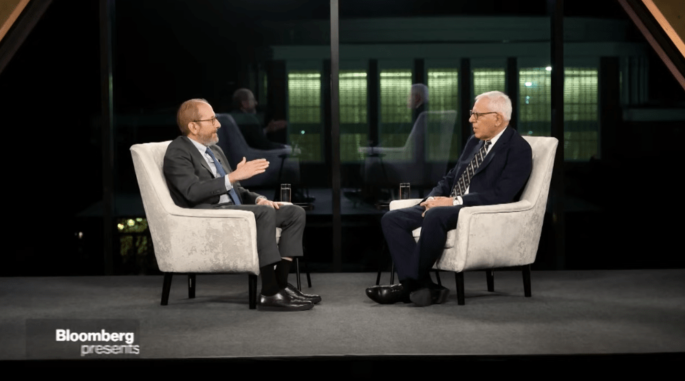 two men sitting in chairs on a stage talking.
