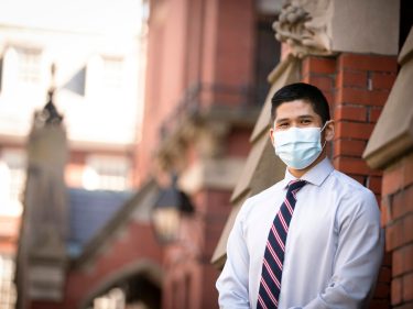 A student wearing a mask stands by a brick building