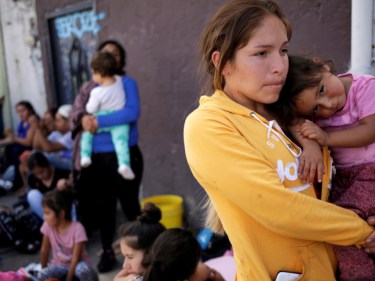 A woman holds a small child while other refugees sit on the ground behind them