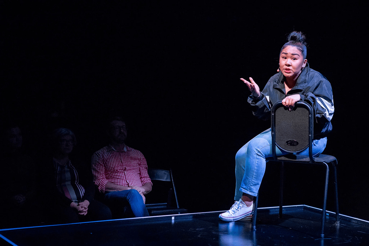 A young woman delivers a monologue while sitting in a chair on a stage