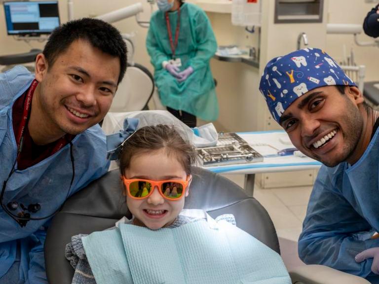 Two student dentists crouch beside a young patient in a dental chair